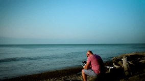 A man is sitting on a pile of driftwood on the Lake Erie coastline flying a drone on a summer day.
