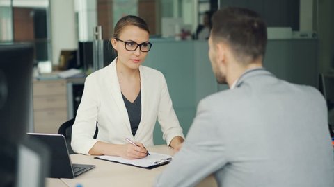 Friendly businesswoman in glasses and suit is interviewing a male candidate for job in office. People are talking asking and answering questions, girl is writing.