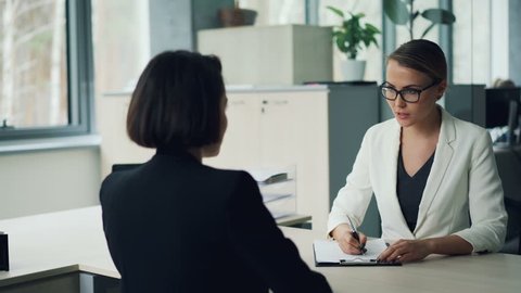 Good-looking young lady from human resources department is interviewing a female professional shaking hands then asking questions and writing. Employment and youth concept.