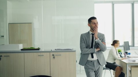 Stressed young man in suit is standing in office waiting for job interview while female interviewer is talking to another candidate. Emotions and employment concept.