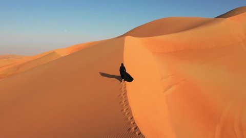 Aerial view from a drone following a young woman in traditional black abaya walking on massive sand dunes in the Empty Quarter desert. Abu Dhabi, UAE.