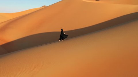 Aerial view from a drone following a young woman in traditional black abaya walking on massive sand dunes in the Empty Quarter desert. Abu Dhabi, UAE.