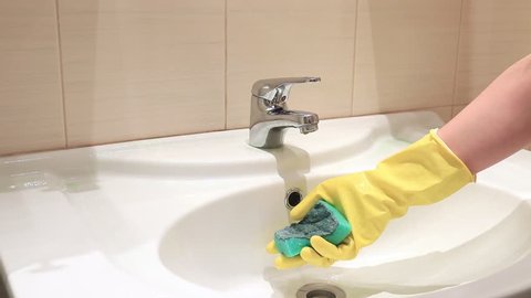 Unrecognizable woman in yellow rubber gloves washing a sponge with water in bathroom sink. Cleaning and washing concept