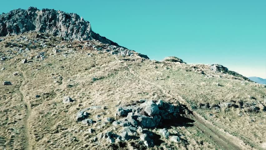 Drone footage towards a steep hill and a rocky out crop with rocks, green grass and walking tracks. Rapaki track Christchurch New Zealand. beautiful blue Skys, rocks and rocky out crops.
