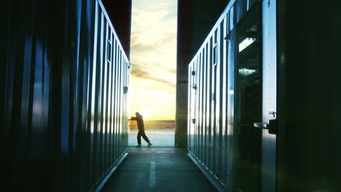 Man Opening Door of a Container Warehouse at Dusk. Orange Sky in the Background. 