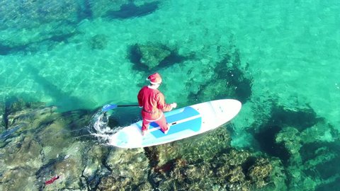 Porto Rafti, Attica / Greece - December 22 2018: Aerial drone video of fit man dressed as Santa Claus practising SUP or Stand Up Paddle in popular beach of Avlaki
