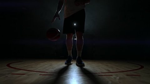 Basketball player in sportswear red shorts and a blue t-shirt goes on a dark basketball court in the backlighting coming out of the smoke knocks a basketball ball on the floor looking at the camera in