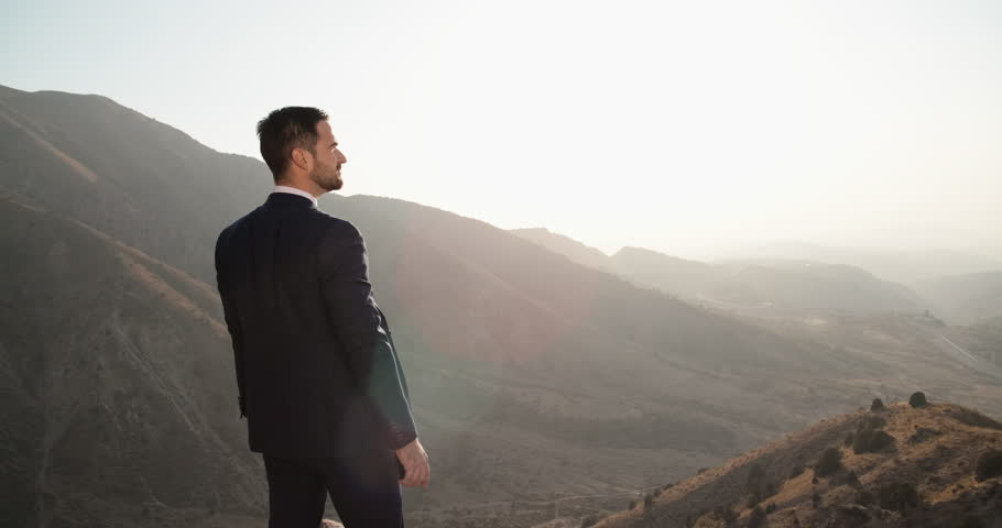Young confident entrepreneur in black suit standing on top of a mountain, looking far at sunset - way to success, on top of the world concept 4k