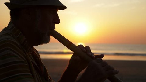 Silhouette of senior man playing bamboo flute on the sandy beach at sunset