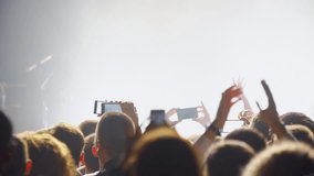 People crowd partying at music concert, jumping and clap their hands against strobing flashing lights, dancing fans, taking photos and recording video with smart phones