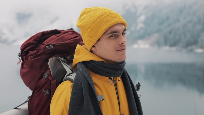 Portrait of a young hiking man with backpack standing standing on nature in the winter, giving thumb up. Beautiful mountains and snow-covered lake background