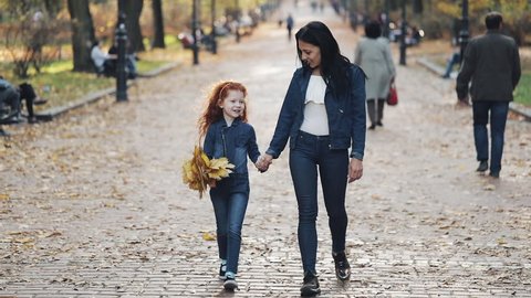 A beautiful mother and her cute little daughter walking in the autumn park. They walk around in the middle of people. Little girl holding yellow leaf bunch in her hand
