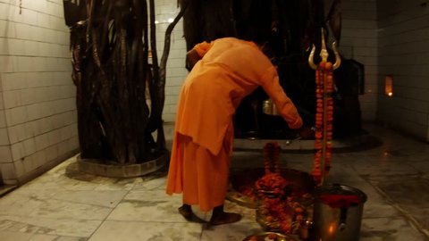 Monk in orange clothes waves with fan near shivlinga decorated with flowers evening hindu ceremony Aarti inside Shiva temple