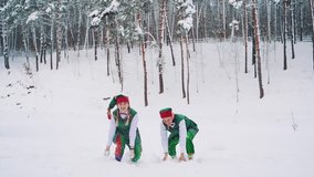 Funny elves in green suits with hats are throwing up snow and looking at it on the background of a winter park. Slow motion