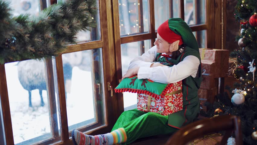 Elf in a green suit sitting on the windowsill with a gift in his hand and a New Year