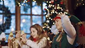 A little girl is sitting at a table with an elf and tearing off a letter with wishes to Santa Claus on the background of the Christmas tree in the room. Festive atmosphere. Blurred Background