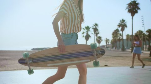Young beautiful female hipster skater long blond hair girl in mini jeans skirt riding and keeping skateboard or longboard in summer hot day on street with palms. Siting and enjoying skate active sport