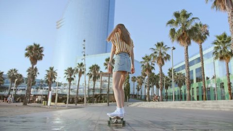 Young beautiful female hipster skater long blond hair girl in mini jeans skirt riding on beach street with palms on skateboard or longboard in summer hot day. Enjoying skateboarding and active sport