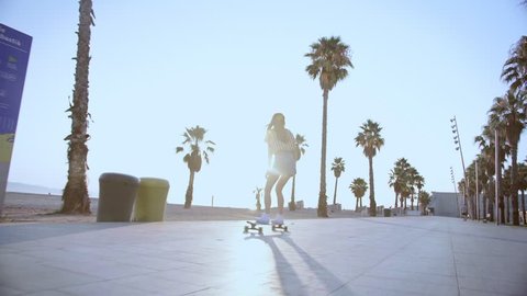 Young beautiful female hipster skater long blond hair girl in mini jeans skirt riding on beach street with palms on skateboard or longboard in summer hot day. Enjoying skateboarding and active sport