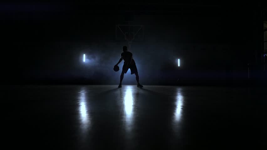A man with a basketball on a dark basketball court against the backdrop of a basketball ring in the smoke shows dribbling skills illuminated by three lanterns in backlight Royalty-Free Stock Footage #1021511815
