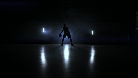 A man with a basketball on a dark basketball court against the backdrop of a basketball ring in the smoke shows dribbling skills illuminated by three lanterns in backlight