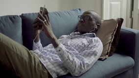 African american man listening music with smartphone on couch. Relaxed young man in earphones lying on sofa and enjoying music. Technology concept