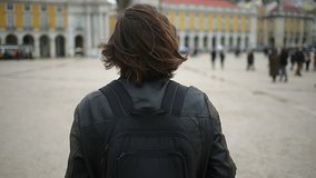 Rear view of man with dark long hair walking in old city. Male tourist with backpack looking around on square. Travel concept