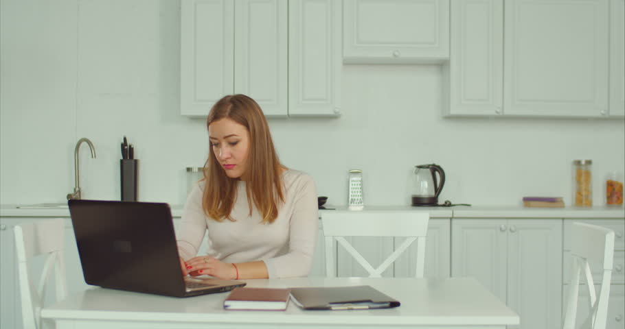 Affectionate caring single mother working laptop, embracing and kissing her cute little daughter with love and happiness in domestic kitchen. Loving freelance mum always has time for her child.