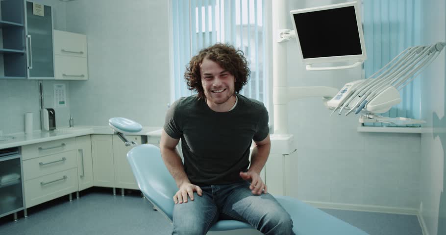 Very charismatic young patient man in a dental clinic room sitting on dentis chair after a teeth check very happy looking in front of the camera and showing a big like. 4k