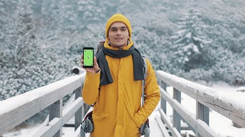 Portrait of a young attractive man in winter clothes showing smartphone with green screen looking into the camera. Chroma Key gadget nature winter