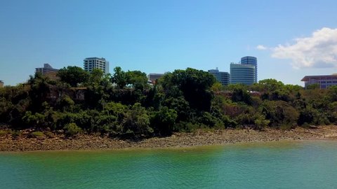 Drone footage flying up and towards Darwin city from a southerly direction over the Darwin harbour revealing Darwin city from behind the foreshore tree line.