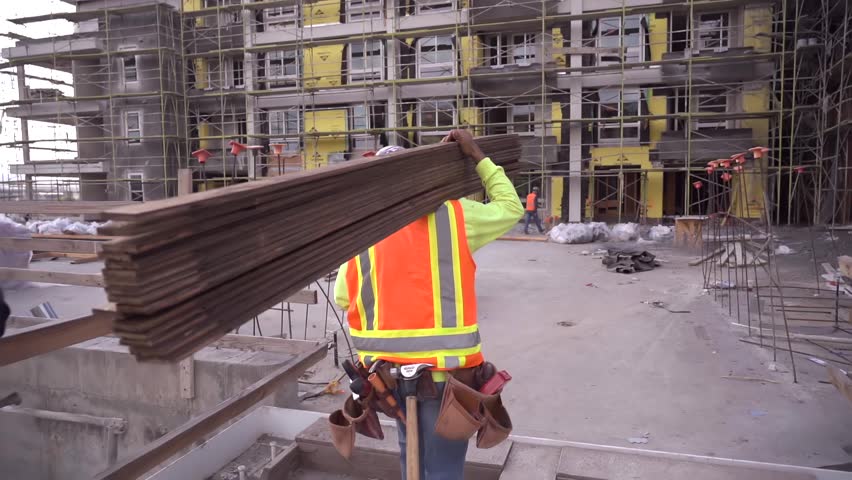 Construction worker walking and working on a construction site wearing a tool belt and high visibility clothing carrying lumber.  Royalty-Free Stock Footage #1021545157