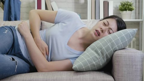 slow motion of young woman with stomach ache lying on couch in apartment. housewife feeling painful suffering from abdomen hurt. sick lady resting at home pain belly hands holding.