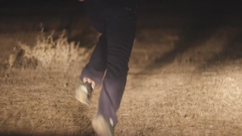 Man running from chasing car. Stock. Closeup of feet of fleeing male in headlights at night in field