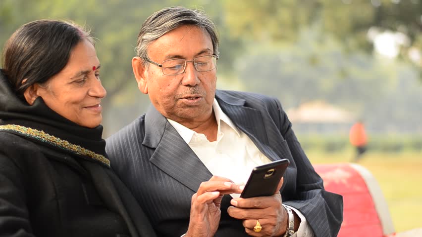 Senior citizen couple reading from / working on a mobile, reading jokes and talking, smiling, laughing  in a park in Delhi, India