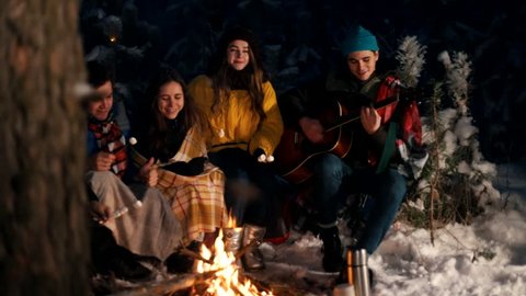 Group of happy friends sitting in winter forest by the fire and eating marshmallows. A young man playing guitar.