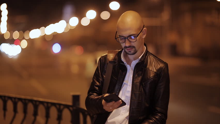 Man sms texting using app on smart phone at night in city. Handsome young business man using smartphone smiling happy wearing suit jacket outdoors. Urban male professional in his 20s.