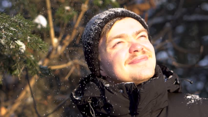 Portrait of young, smiling man with moustache looks up, enjoys snowfall in winter forest, squints his eyes from the bright sun. Handsome, happy man likes falling snow over his head in a sunny day.