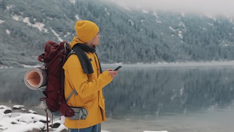 American traveler man using smartphone in hiking winter tour. Snowy lake and mountains against background. Traveling and communication concept: hiker scrolling and tapping on line by cell phone