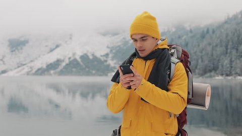 Young traveler man using smartphone in hiking winter tour. Snowy blurred mountains cape and lake. Traveling and communication concept: hiker scrolling and tapping on line by cell phone