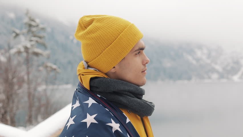 Traveller man with flag of America standing in the snow-covered mountains near beautiful lake. Hiker looking at the beautiful nature