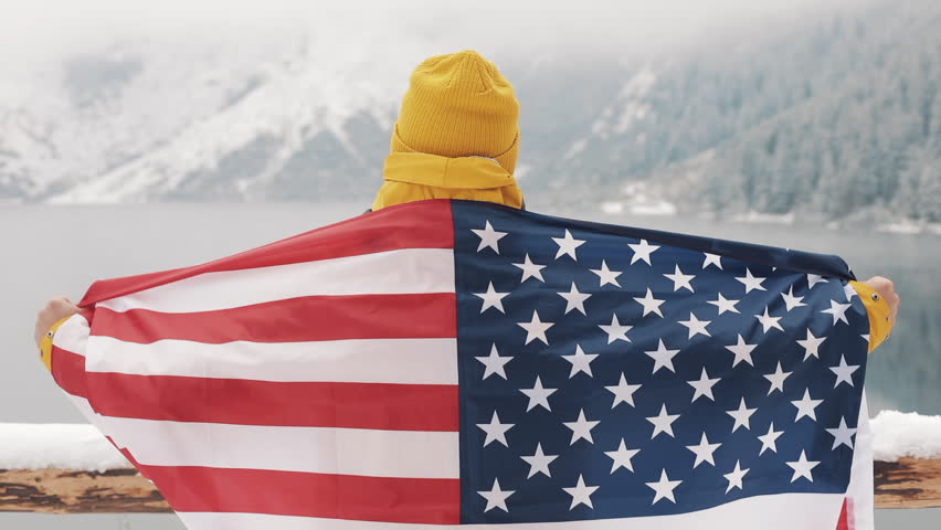 Traveller man with flag of America standing in the snow-covered mountains near beautiful lake. Hiker looking at the beautiful nature