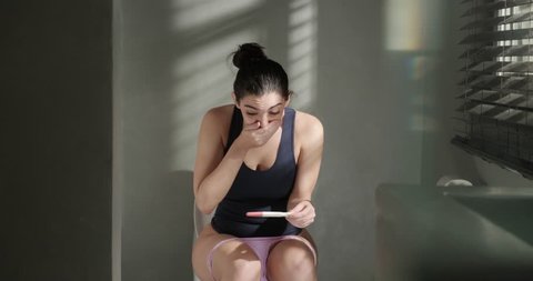 Anxious young white woman with nervous feelings on bathroom. Caucasian girl holding a positive pregnancy test, crying after finding result. Pregnancy test kit in hands. Emotional female in restroom