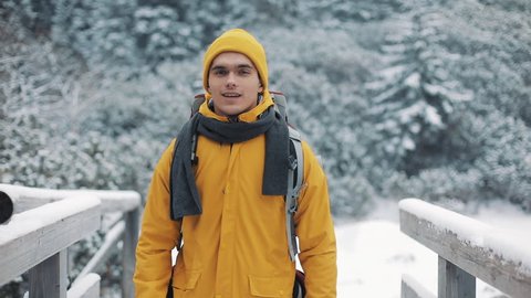 Traveling in the mountains. Young man wearing yellow winter clothing walking on the mountain covered with snow. He goes on a bridge across the river and looks at the camera. Trip, adventure, climbing