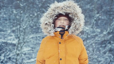 Boy dressed in Warm Hooded Casual Parka Jacket Outerwear looking into camera and clapping with snow full palms with falling snow background. Outdoor time and winter outfit concept video.