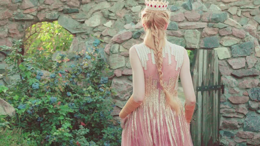 Medieval fantasy woman blonde queen, walks near castle, opens  wooden door and goes out into nature from park. Luxurious pink royal Victorian vintage dress. Portrait happy girl princess, cheerful face