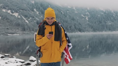 American traveler man using smartphone in hiking winter tour. Snowy lake and mountains against background. Traveling and communication concept: hiker scrolling and tapping on line by cell phone