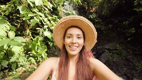 Young Mixed Race Tourist Blogger Girl in White Dress and Straw Hat Making Selfie Video and Talking on Mobile Phone with Amazing Wild Jungle Waterfall on Background. Lifestyle Travel 4K Footage. Bali,
