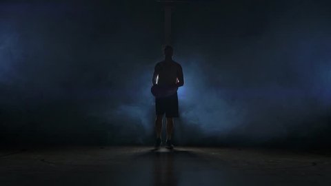 Basketball player goes straight to the camera in a dark room with a backlit back in the smoke looking at the camera in slow motion. Steadicam