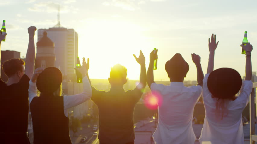 Rear view of group of young men and women holding beer bottles and holding hands up in the air while jumping and dancing on urban rooftop at sunset | Shutterstock HD Video #1021627894